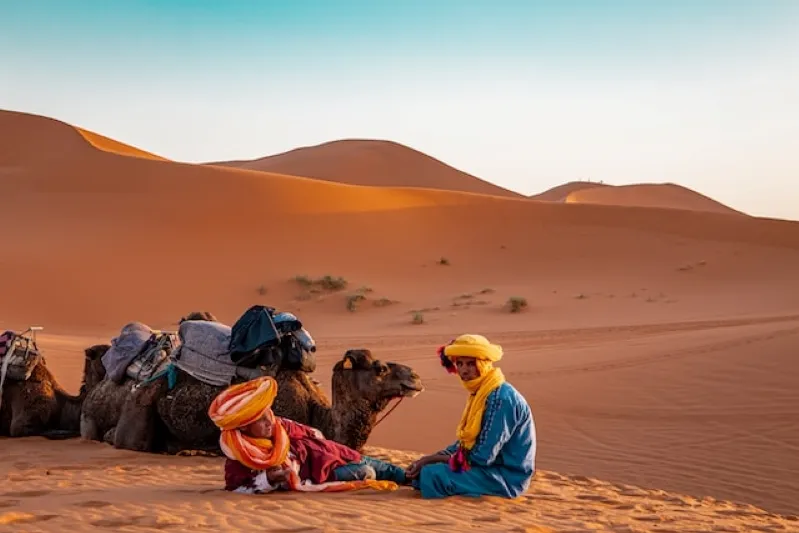 Two individuals in vibrant attire seated on the sand beside camels, showcasing a lively scene in Morocco's landscape.