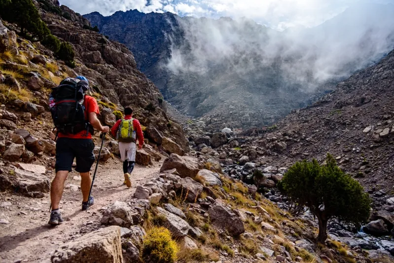 Two hikers with backpacks ascend a mountain trail in Morocco, surrounded by stunning natural scenery. 