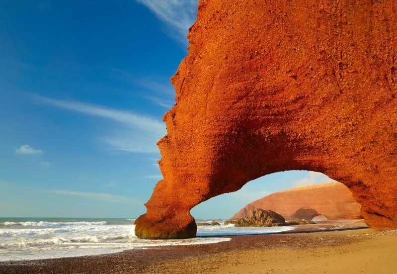A stunning red rock arch stands majestically at a Moroccan beach, surrounded by the serene desert landscape. 