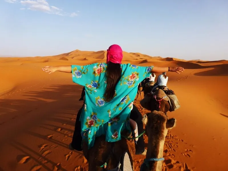 A woman in a vibrant dress rides a camel through the Moroccan desert, showcasing the beauty and safety of the region. 