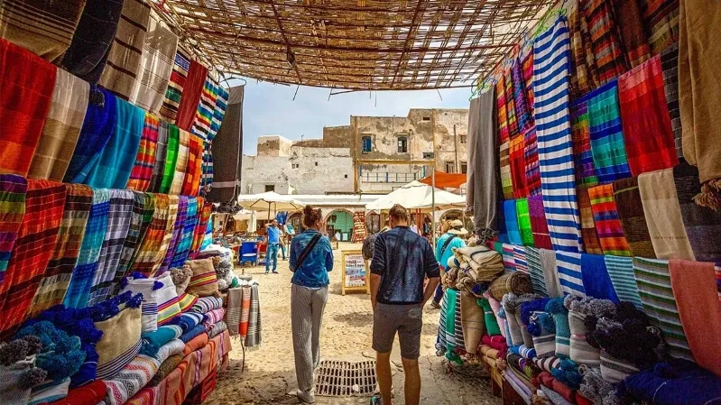 A vibrant market scene in Morocco, where people stroll among colorful blankets and textiles, showcasing local craftsmanship. 