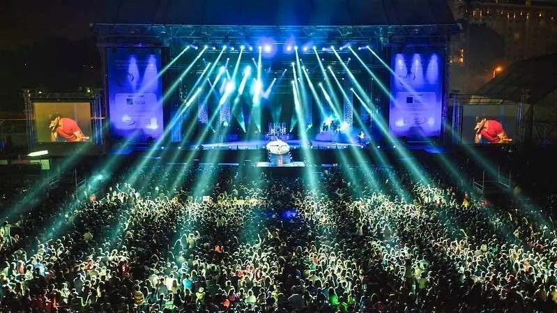 A vibrant concert crowd illuminated by bright lights, set against a backdrop of stunning moroccan architecture.