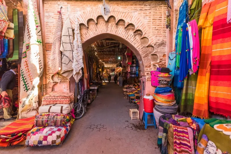 A vibrant narrow alley adorned with colorful blankets and pillows, showcasing Moroccan textiles and culture.