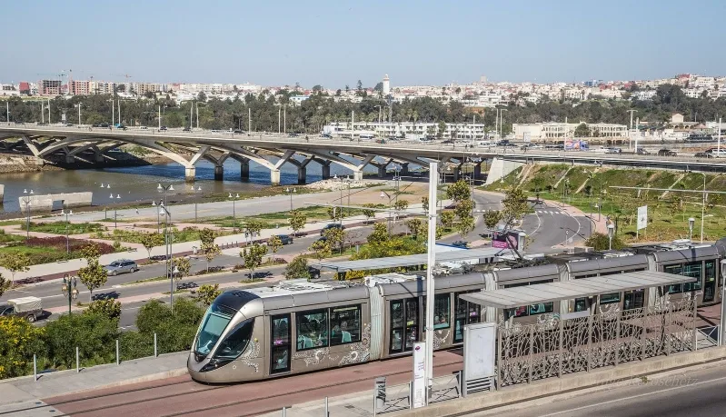 A train moves along the tracks, passing under a bridge in a scenic landscape, showcasing Morocco's transportation infrastructure.