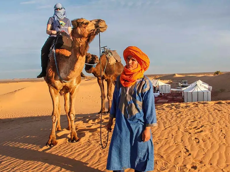 A man in an orange turban and blue dress stands beside two camels in morocco sahara desert tours from marrakech.