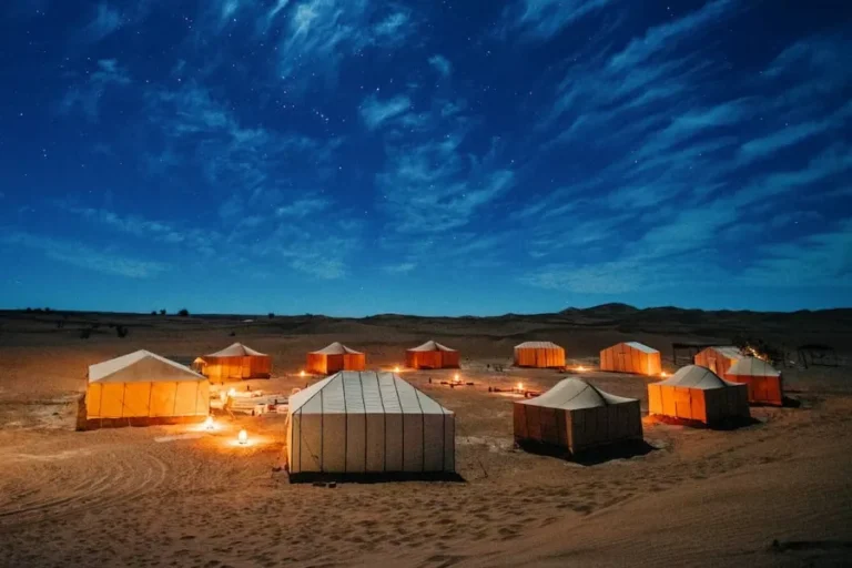 A serene Moroccan desert camp at night, featuring tents illuminated by a warm fire under a starry sky.