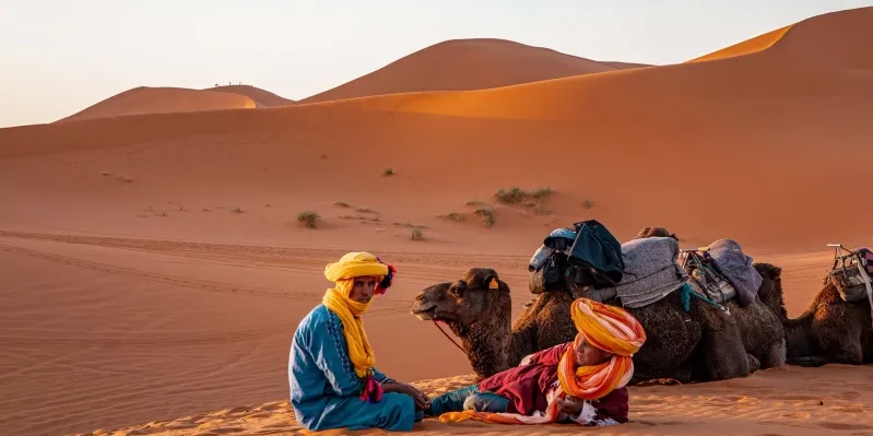 Two individuals in vibrant attire seated on the sand of the Moroccan Sahara Desert, accompanied by camels.
