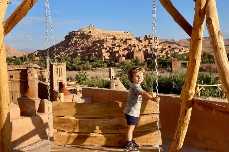 A young boy swings joyfully in front of a vast desert landscape during a day trips from Ouarzazate.