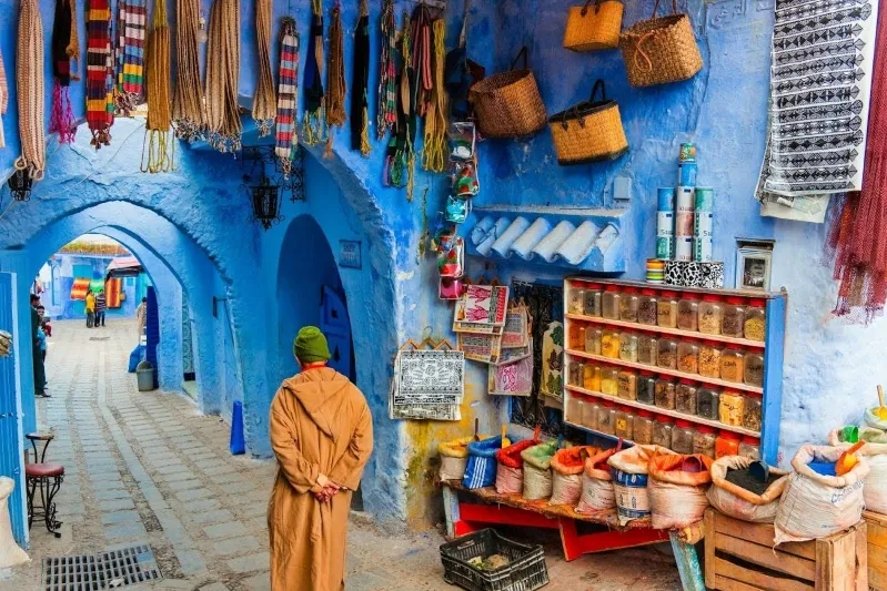 A man in a blue robe walks through a narrow alleyway, showcasing the vibrant culture of Morocco for a travel agency.