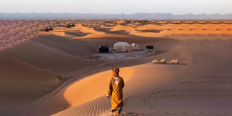 A man walks through a desert landscape, with a tent set up nearby, highlighting the arid environment of Ouarzazate, Morocco.