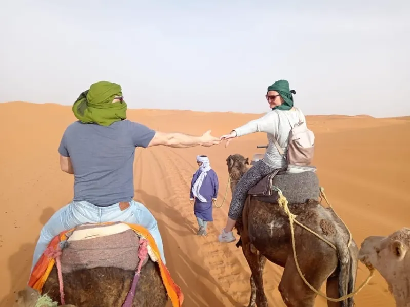 Two individuals on camels in the desert, one holding hands with the other, showcasing a day trip from Ouarzazate.