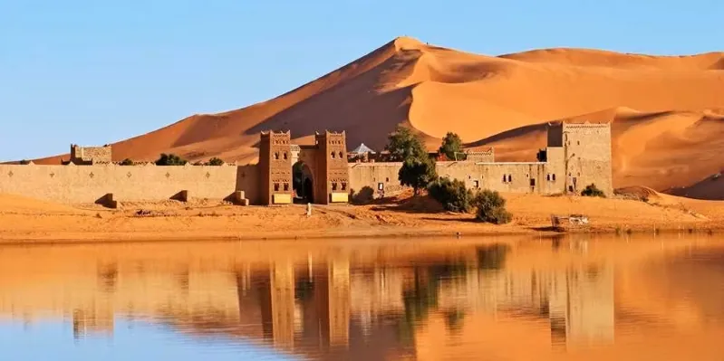 A desert landscape in Ouarzazate, Morocco, featuring sand dunes and a body of water in the background. 