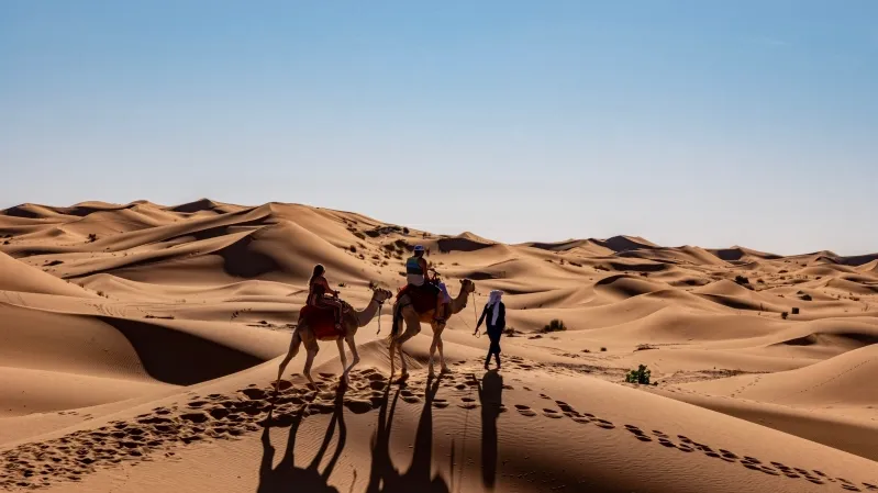  Two individuals riding camels across a vast desert landscape during a Morocco camel tours.