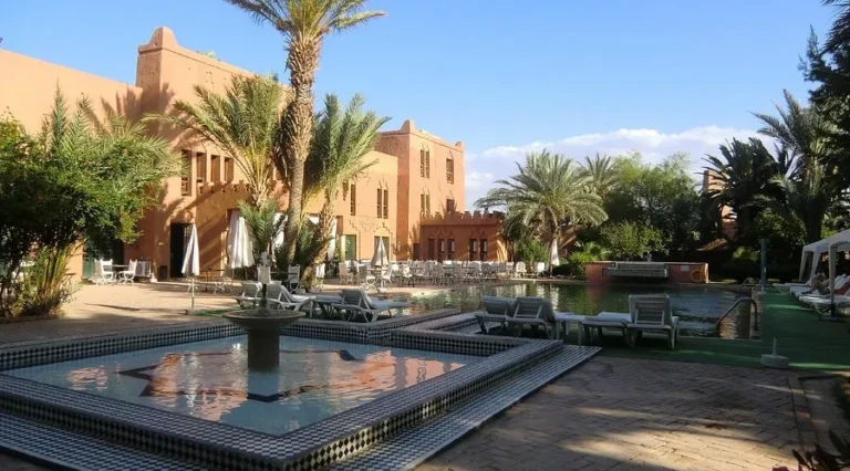 A large pool encircled by palm trees, with a riad building in the background, showcasing a serene oasis in Ouarzazate.