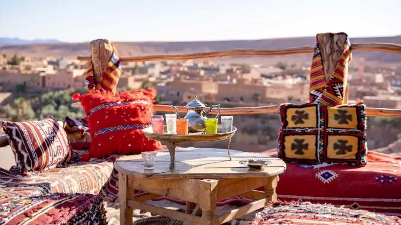 A table with colorful pillows set on a mountain peak, showcasing a scenic view, promoting a vacation in Morocco. 