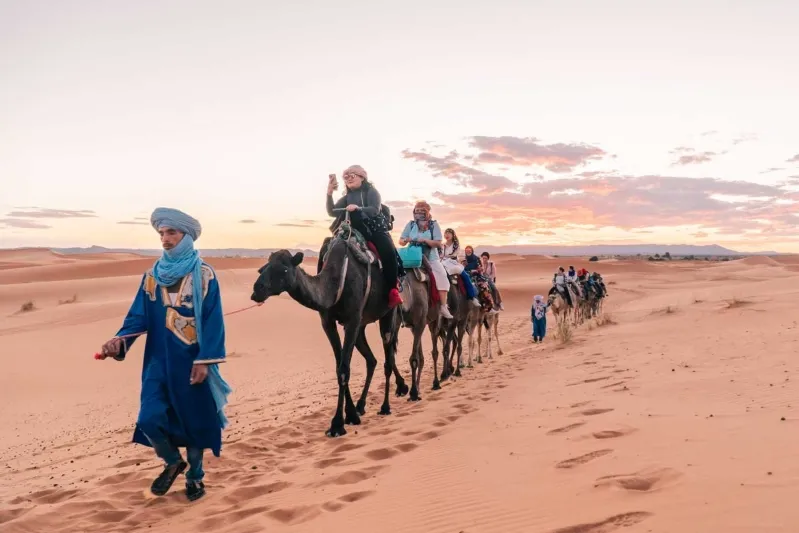 Tourists in blue attire and turbans ride camels across the Moroccan desert, enjoying a scenic adventure.