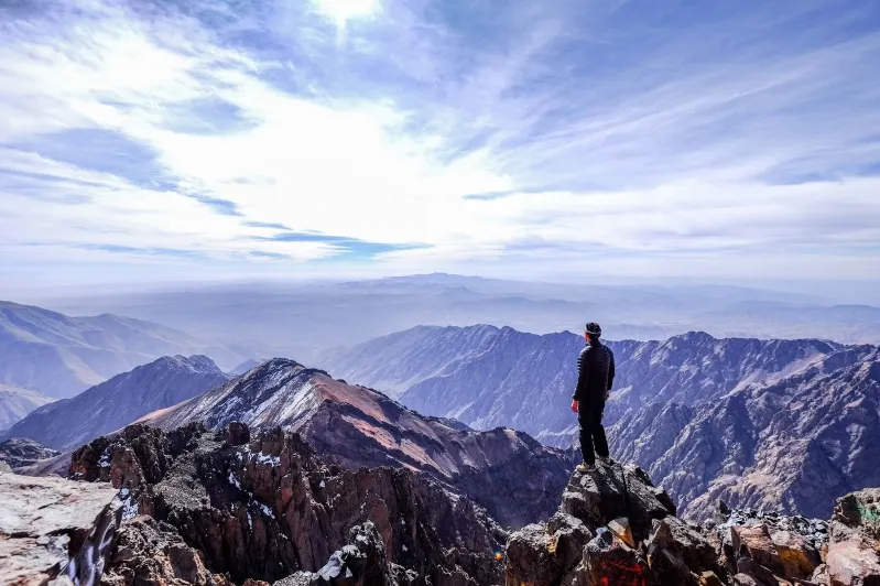 A man stands atop a peak in the Atlas Mountains range, gazing out over the expansive mountain range below.