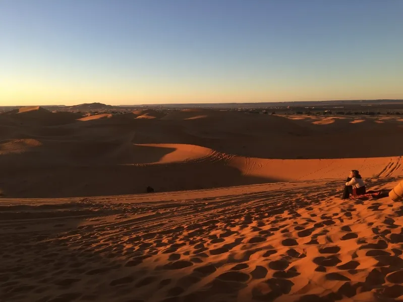 A traveler seated on golden sand dunes at sunset, enjoying the picturesque view, ideal for Morocco tour packages.