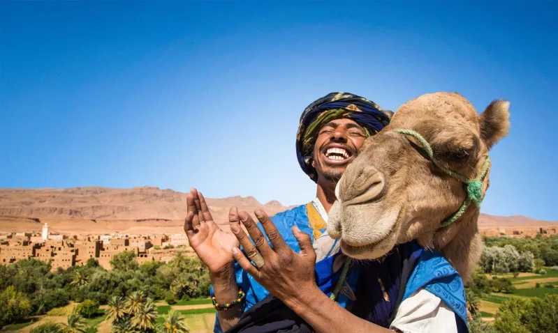 A man wearing a turban and blue attire stands with a camel, representing a moment from a Morocco group tour.
