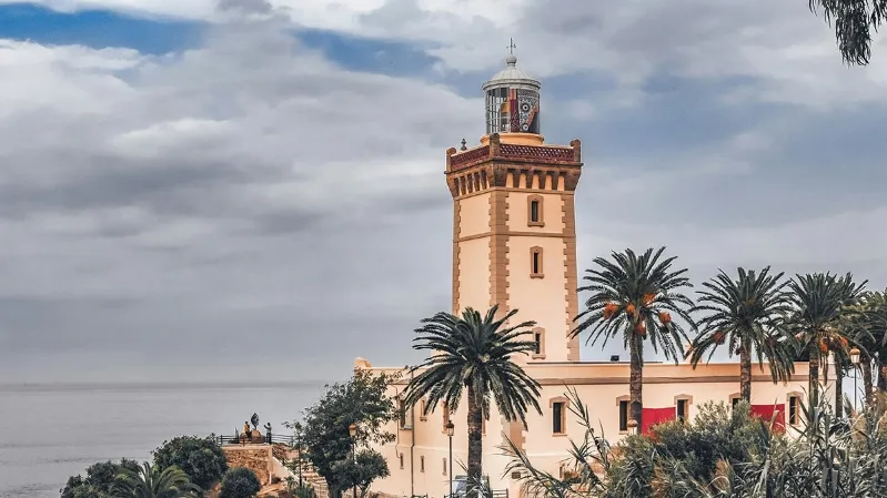 A coastal scene featuring a tower, palm trees, and a lighthouse beside the ocean in Tangier, Morocco.