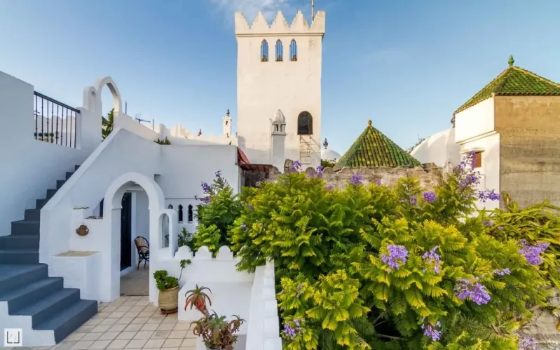 A white house featuring a tower and stairs, set in a Moroccan context with local people nearby.