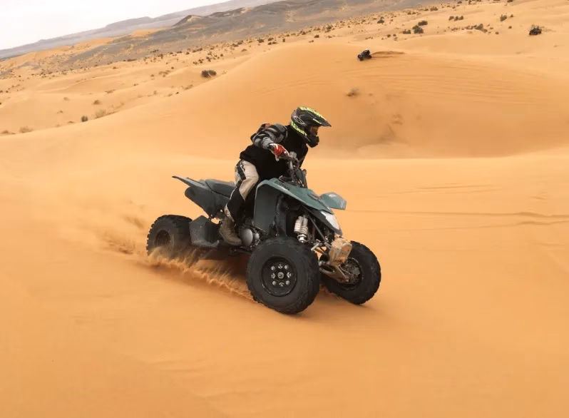A travel agent in Morocco rides an ATV across a sandy desert landscape under a clear blue sky.
